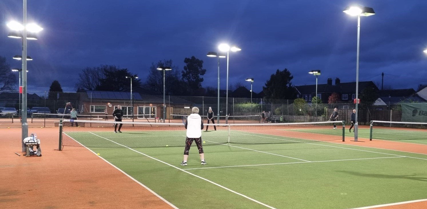 Players on a green tennis court at night time, with floodlights illuminating the courts