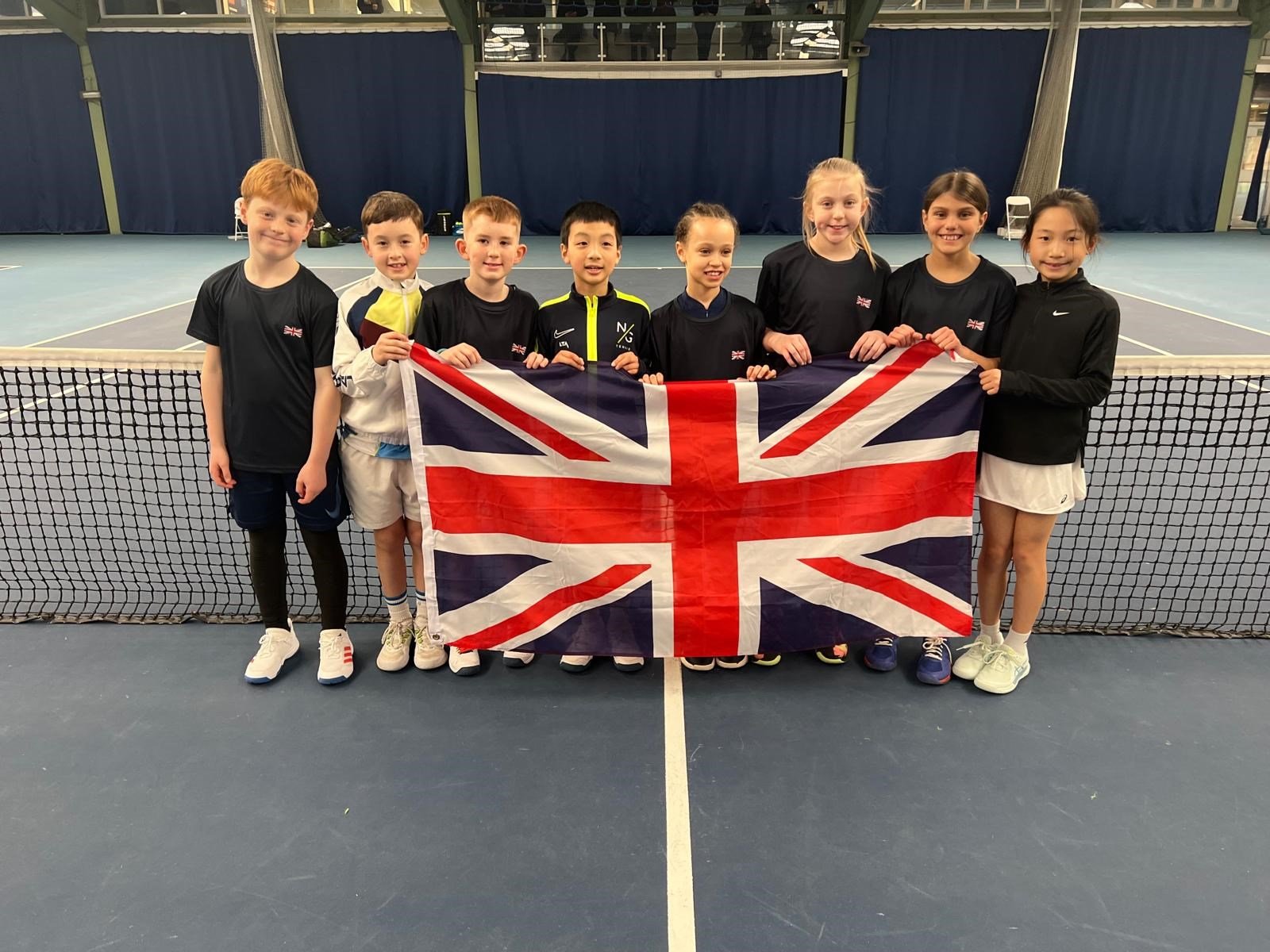 A group of eight 11U players posing by the net on a tennis court, holding up a Union Jack flag in front of them