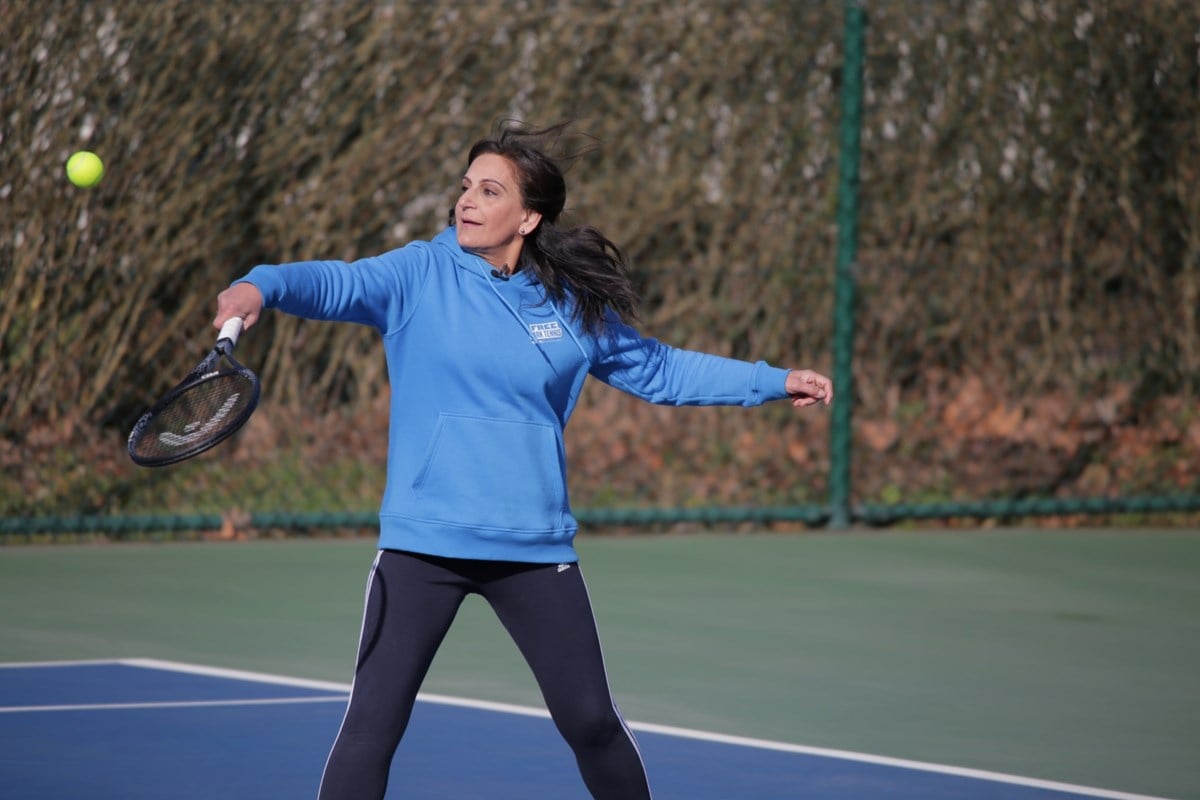 A woman in a blue shirt prepares to strike a tennis ball with her racket on a sunny court.