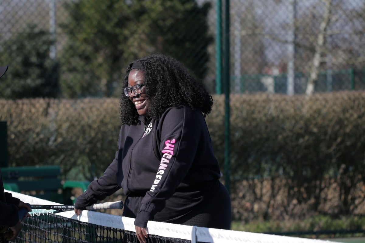 A woman, smiling, stands at a tennis net with a racket in her right hand resting on top of the net. The hands and racket handle of another player are visible on the other side of the net.