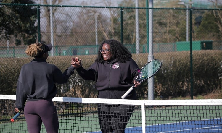 Two women shake hands over the top of a tennis net, each holding a tennis racket in their left hand. Tennis court markings and fecing are visible in the background.