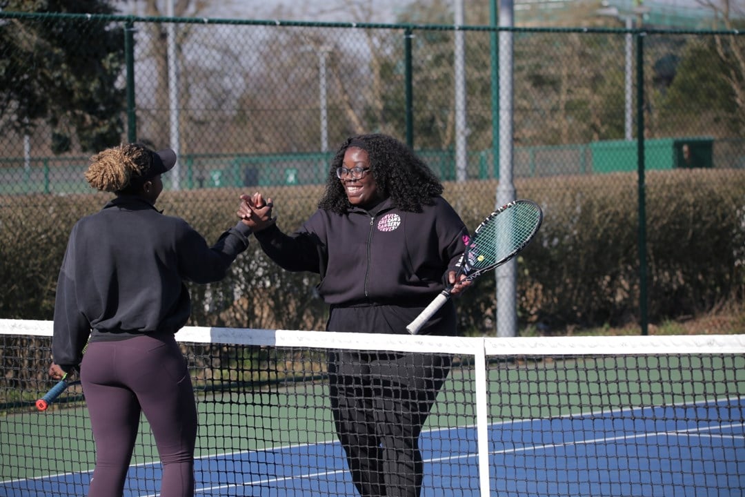 Two women shake hands over the top of a tennis net, each holding a tennis racket in their left hand. Tennis court markings and fecing are visible in the background.