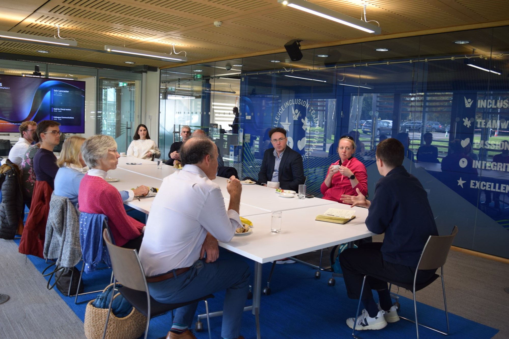 A group of people sat around a white table, with a man, facing away from the camera talking to the wider group