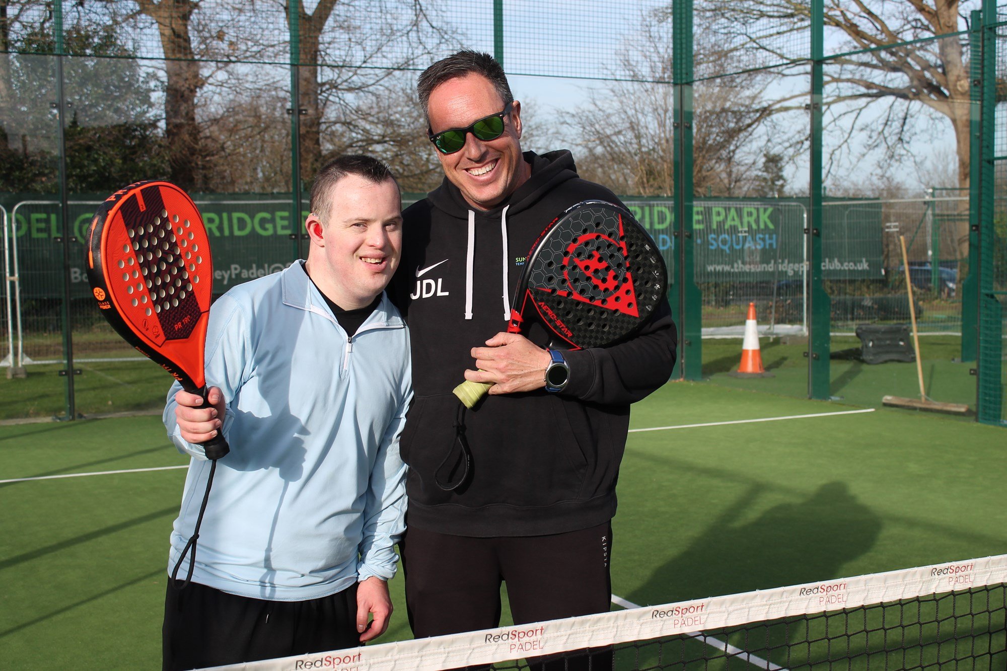 Two men standing side by side, both holding padel bats. One man is wearing all back and is smiling, while the other, standing on the left and also smiling, is wearing a blue top