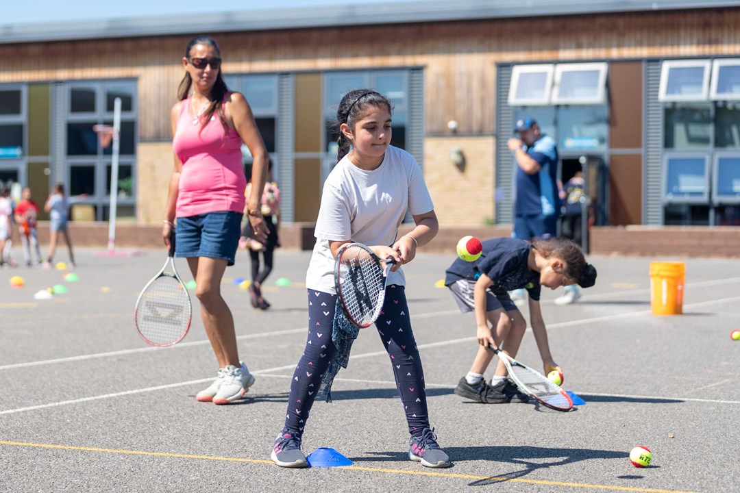 an image of a young girl holding a tennis racket and hitting a ball on court with other children and an older woman stood around her