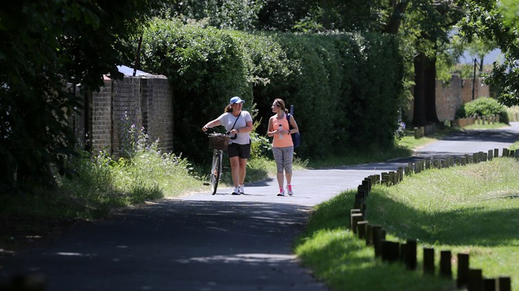 Two women walking on a tarmac path through a park, dressed in sports gear, with the lady on the left guiding a bicycle