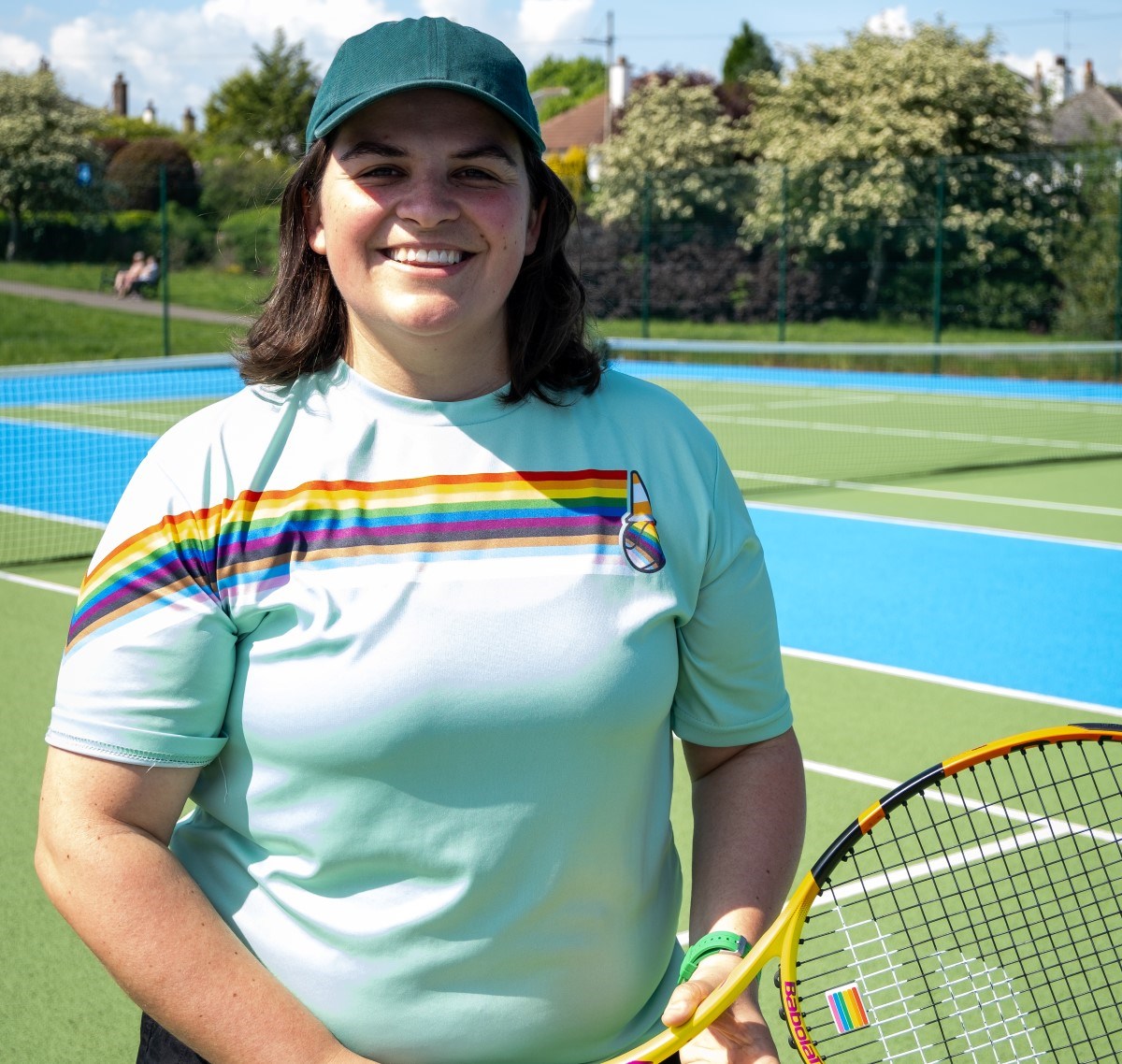 A woman stood on a tennis court smiling at the camera, wearing a blue t-shirt with a rainbow across the front, holding a yellow tennis racket in her hands