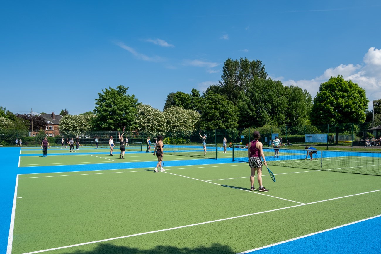 A group of people playing tennis on tennis courts. There are trees in the background and a blue, almost cloudless sky