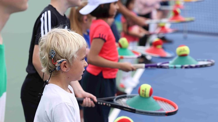 A group of children wearing hearing aids and holding tennis rackets and balls