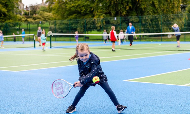 A young girl preparing to hit a ball with her tennis racket surrounded by other children on a park tennis court