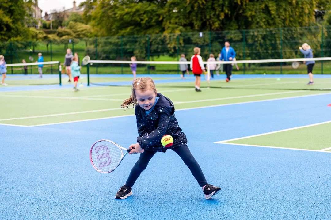 A young girl preparing to hit a ball with her tennis racket surrounded by other children on a park tennis court