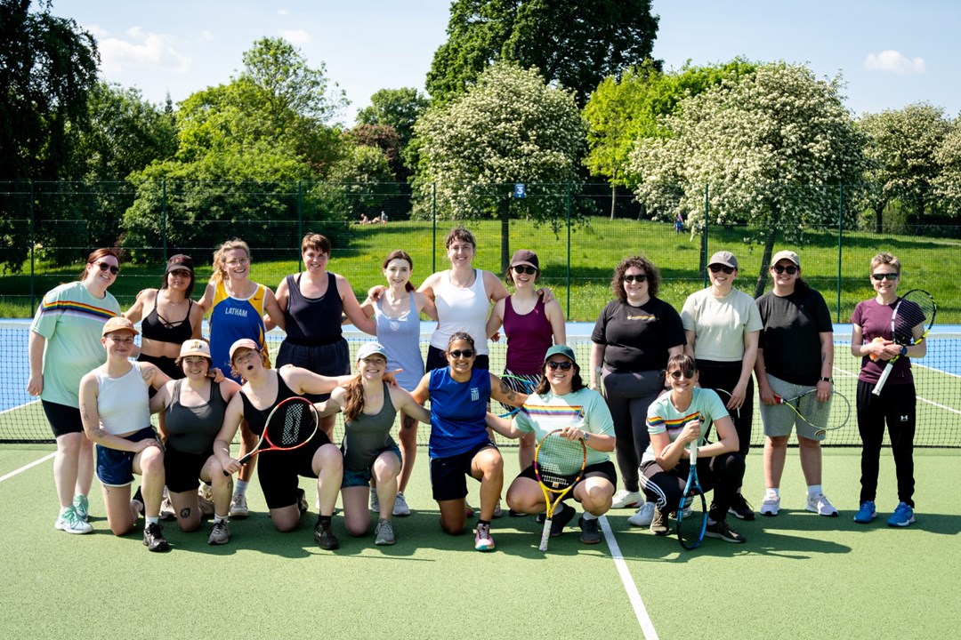 A group of woman stood together on a tennis court, smiling at the camera, with blue sky and greenery in the background