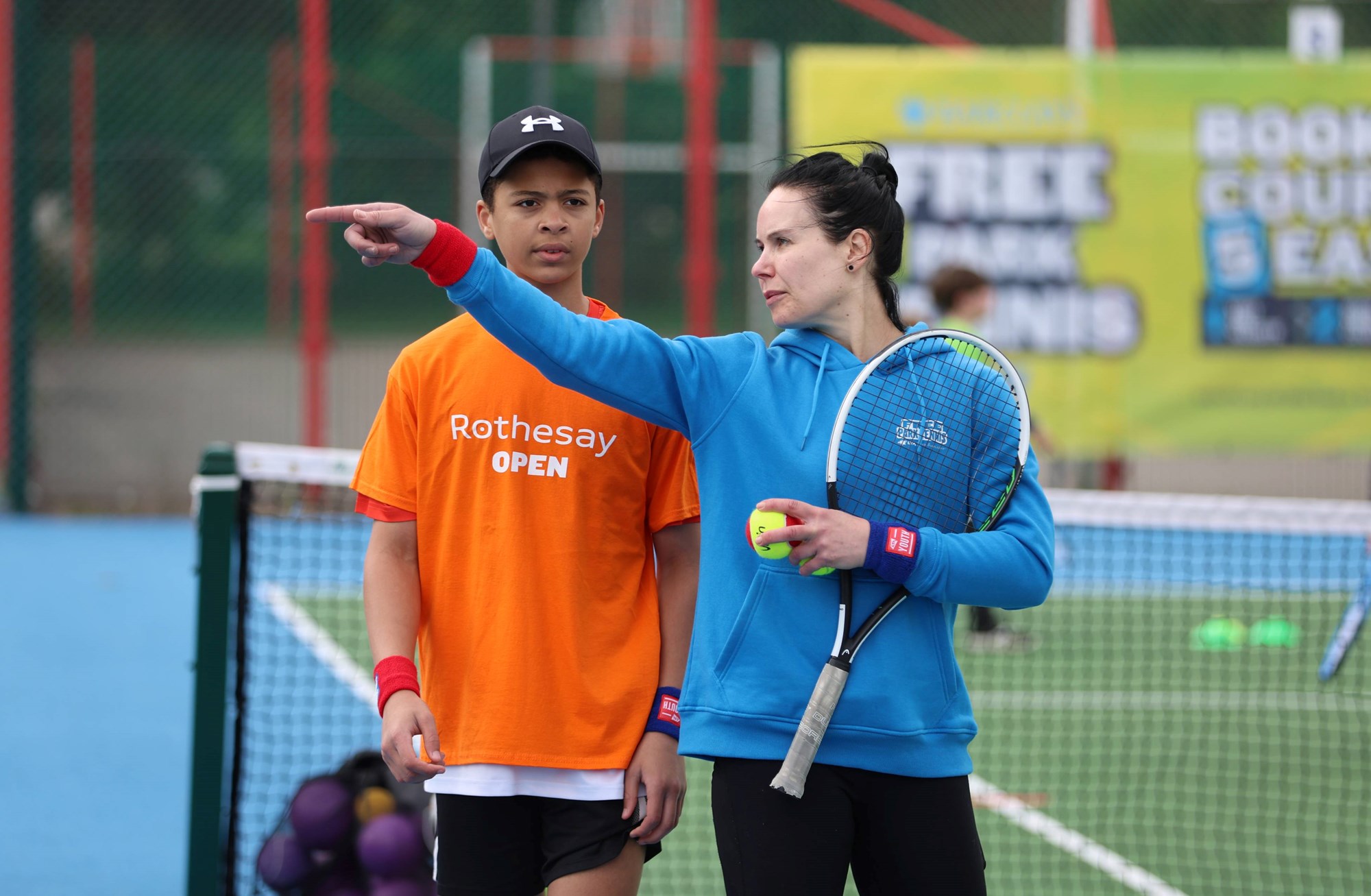 A Free Parks Tennis Activator holding a tennis racket and ball while talking to a young boy wearing a Rothesay Open t-shirt