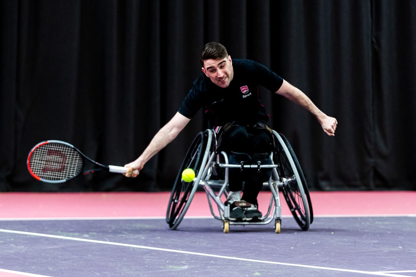 A man in a wheelchair leans forward to try and hit a tennis ball with a racket