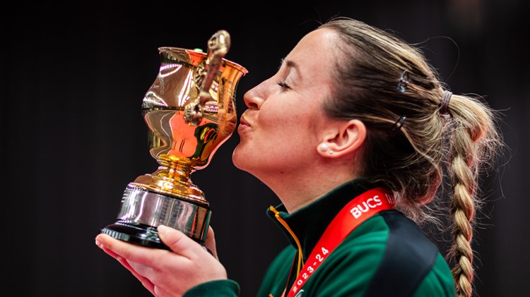 A woman, wearing a green jumper, viewed from the side as she kisses a small trophy and wears a red ribbon around her neck, supporting a medal out of view