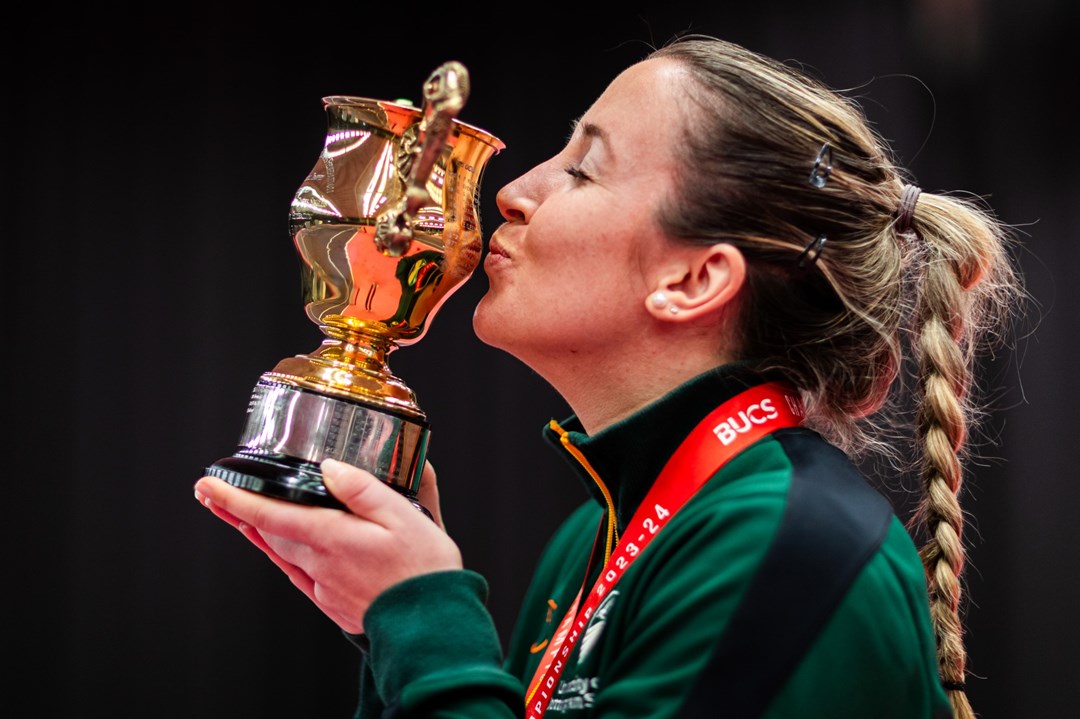 A woman, wearing a green jumper, viewed from the side as she kisses a small trophy and wears a red ribbon around her neck, supporting a medal out of view