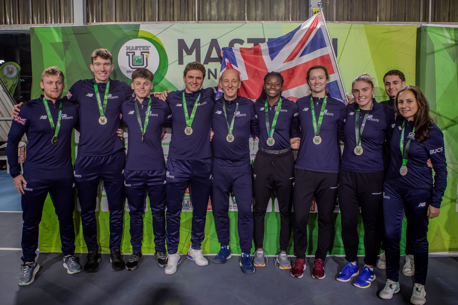 A group of people dressed in dark blue tracksuits stood arm-in-arm with medal around their necks. There is a British flag on the wall behind them