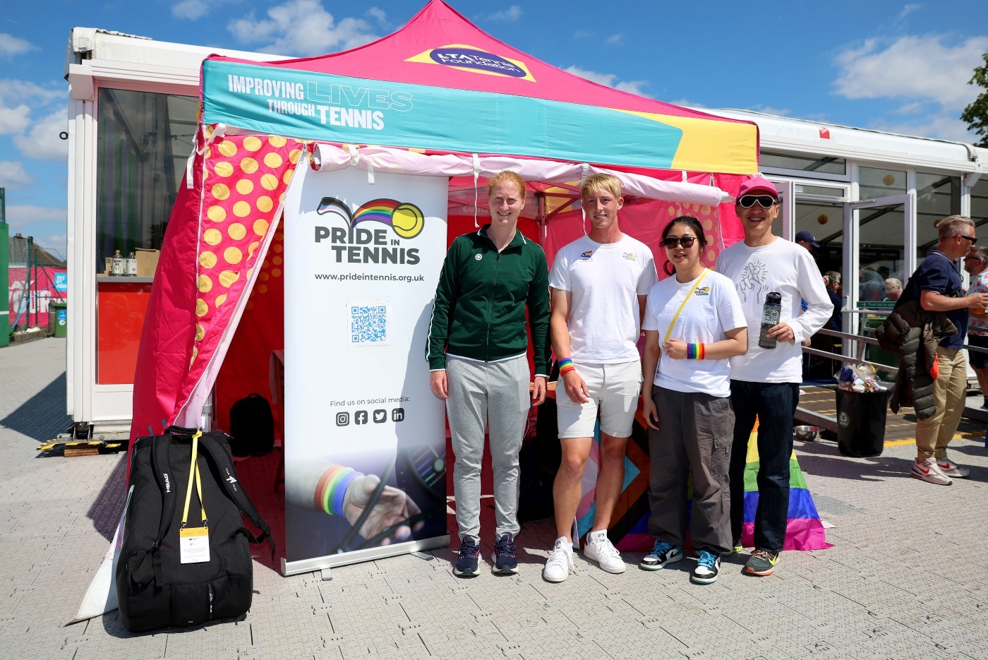 A group of four people stood in front of a large, pink, open-sided gazebo. Three are dressed in white and the fourth, stood on the far left of the group, is dressed in a green tracksuit top and grey tracksuit bottoms. The words, Pride in Tennis, are on a banner next to the group.