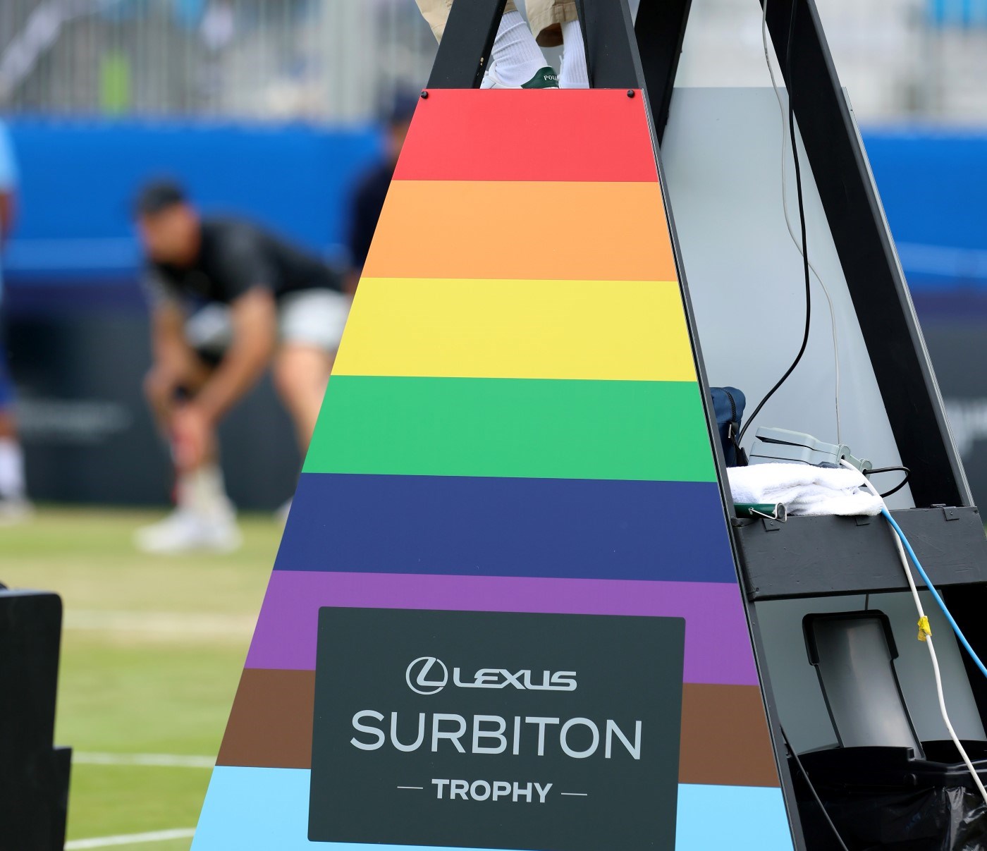 A board with rainbow colours on it with the words, Lexus Surbiton Trophy, in the middle of the board. The board is on the side of a tennis umpire chair, with the shoes of the umpire just visible in the top of the shot