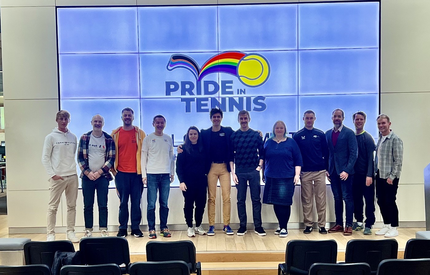 A group of people stood smiling at the camera, with a large screen behind them displaying the words, Pride in Tennis