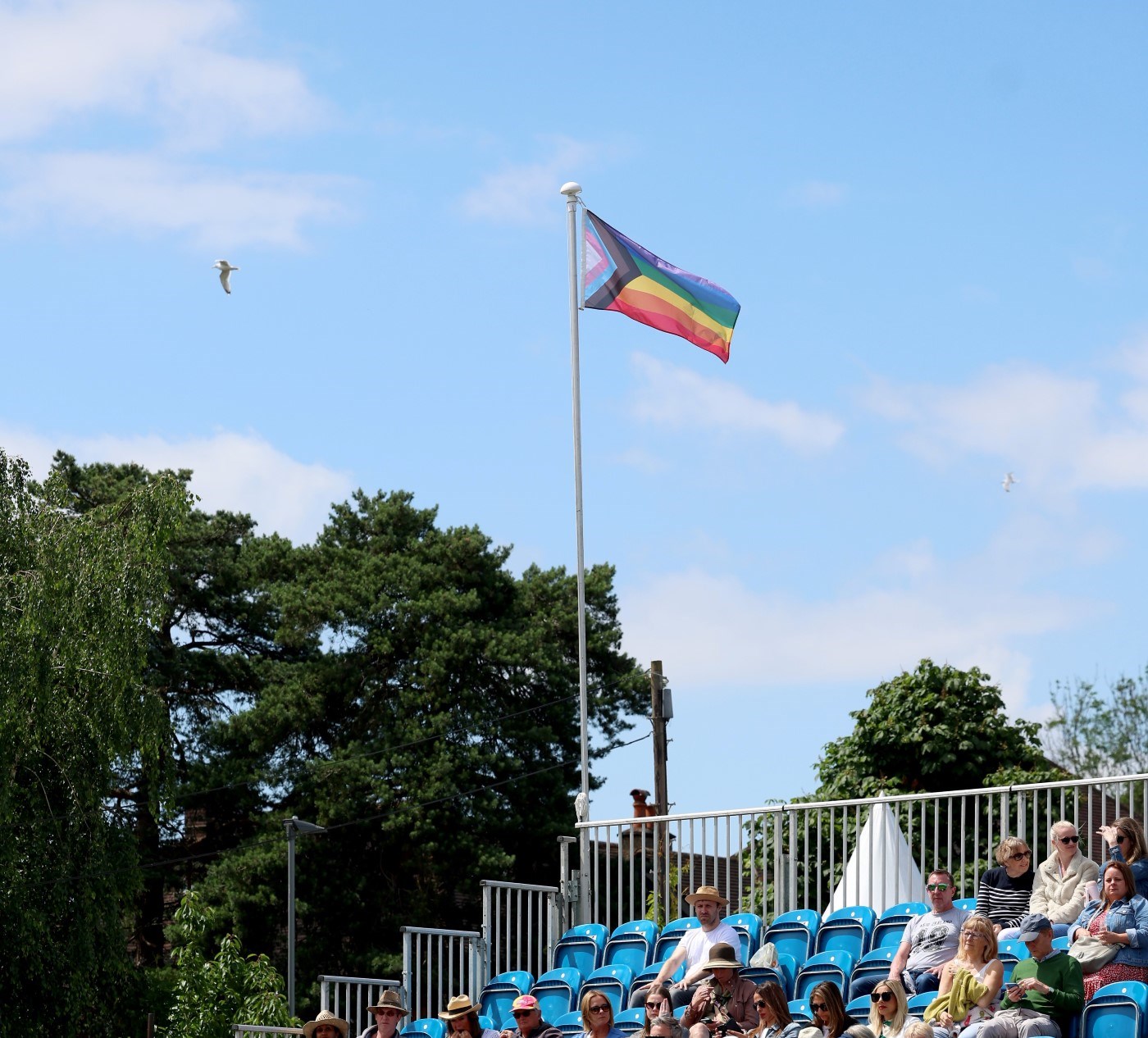 A Progress Pride flag flies above a number of blue seats, with a number of people sitting below the flag. The flag has a variety of colours, with bands running from bottom to top of red, orange, yellow, green, blue and purple, and arrow shaped bands of colour cross-cutting the horizontal bands, running left to right of white, pink, light blue, brown and black