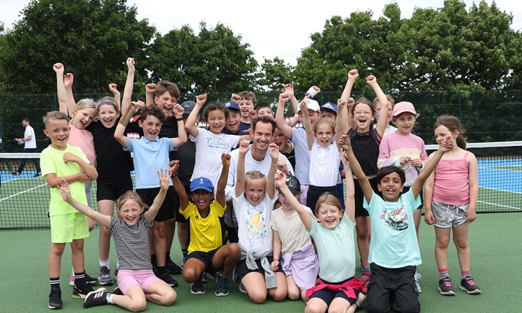 Andy Murray smiling in the middle of a group of young kids cheering with their hands in the air on a park court
