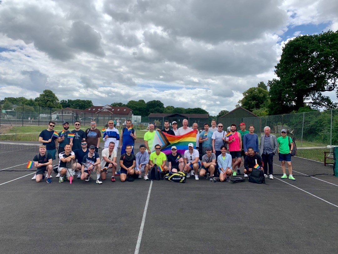 Members of the ACE Players Tennis Croydon LGBTQ+ Tennis Group pictured as group on court, holding the rainbow flag.