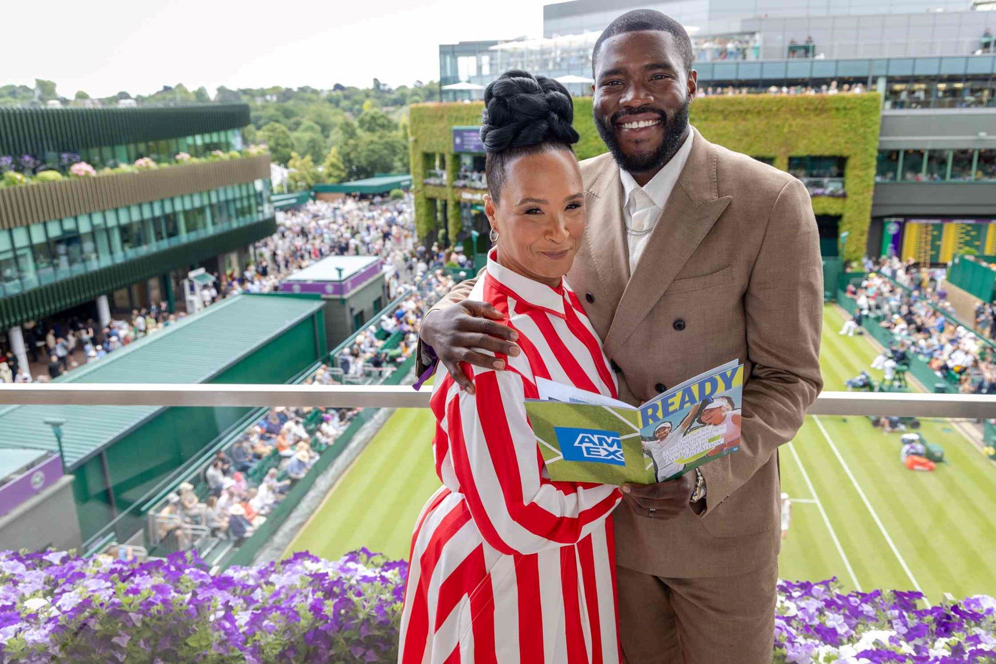 Golda Rosheuvei and Martins Imhangbe smiling on a balcony at Wimbledon while holding a tennis magazine