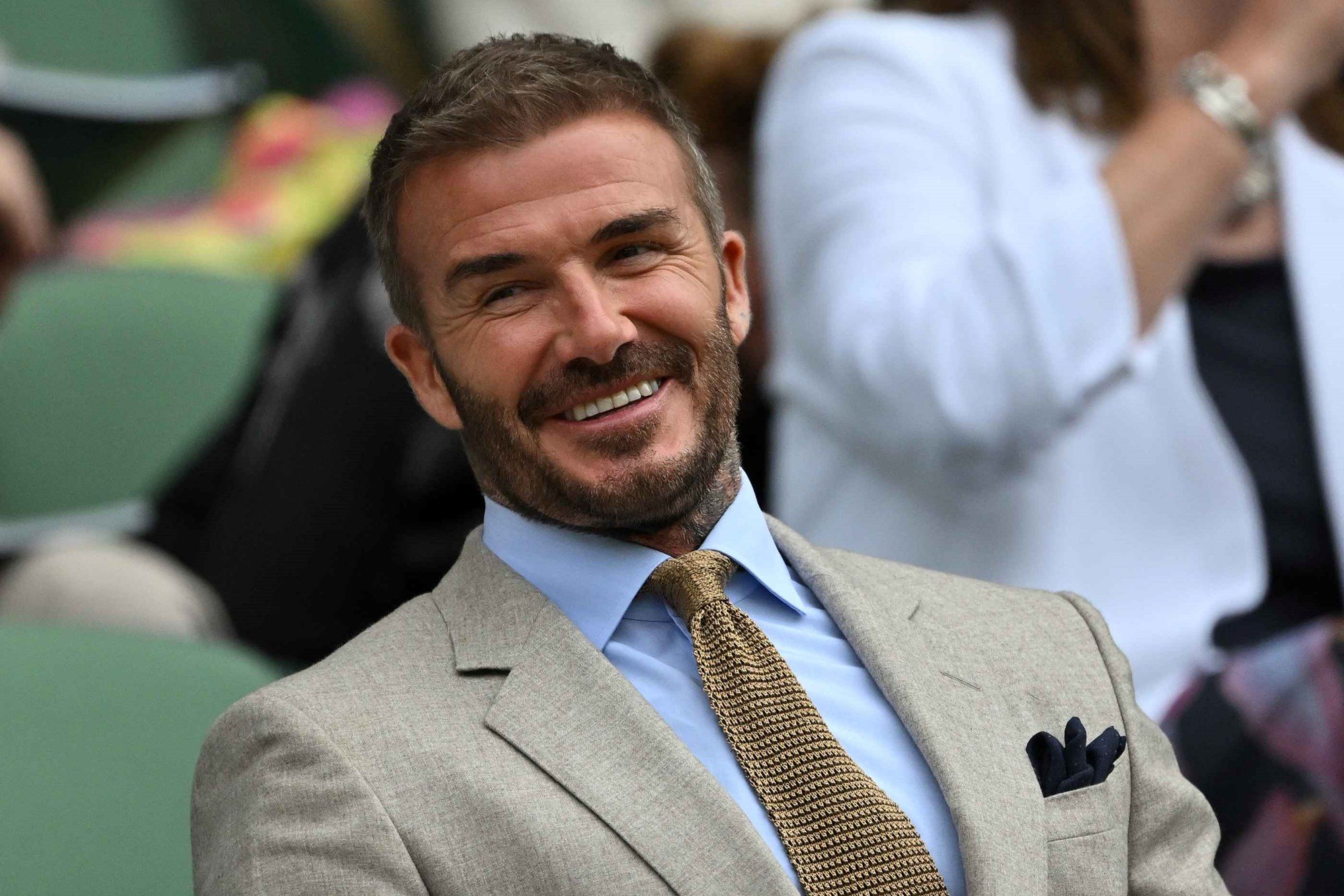 David Beckham wearing a suit and smiling while sat in a seat at Wimbledon