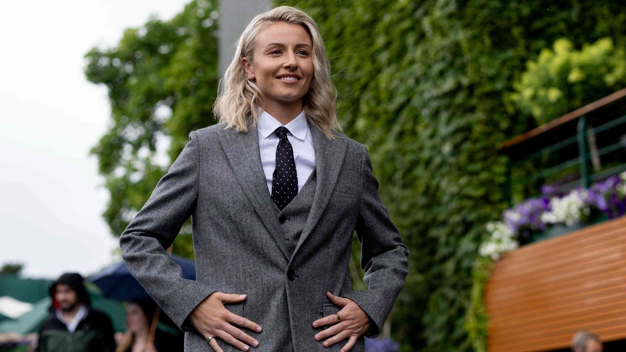 Leah Williamson smiling while wearing a grey suit in front of a wall of flowers at Wimbledon