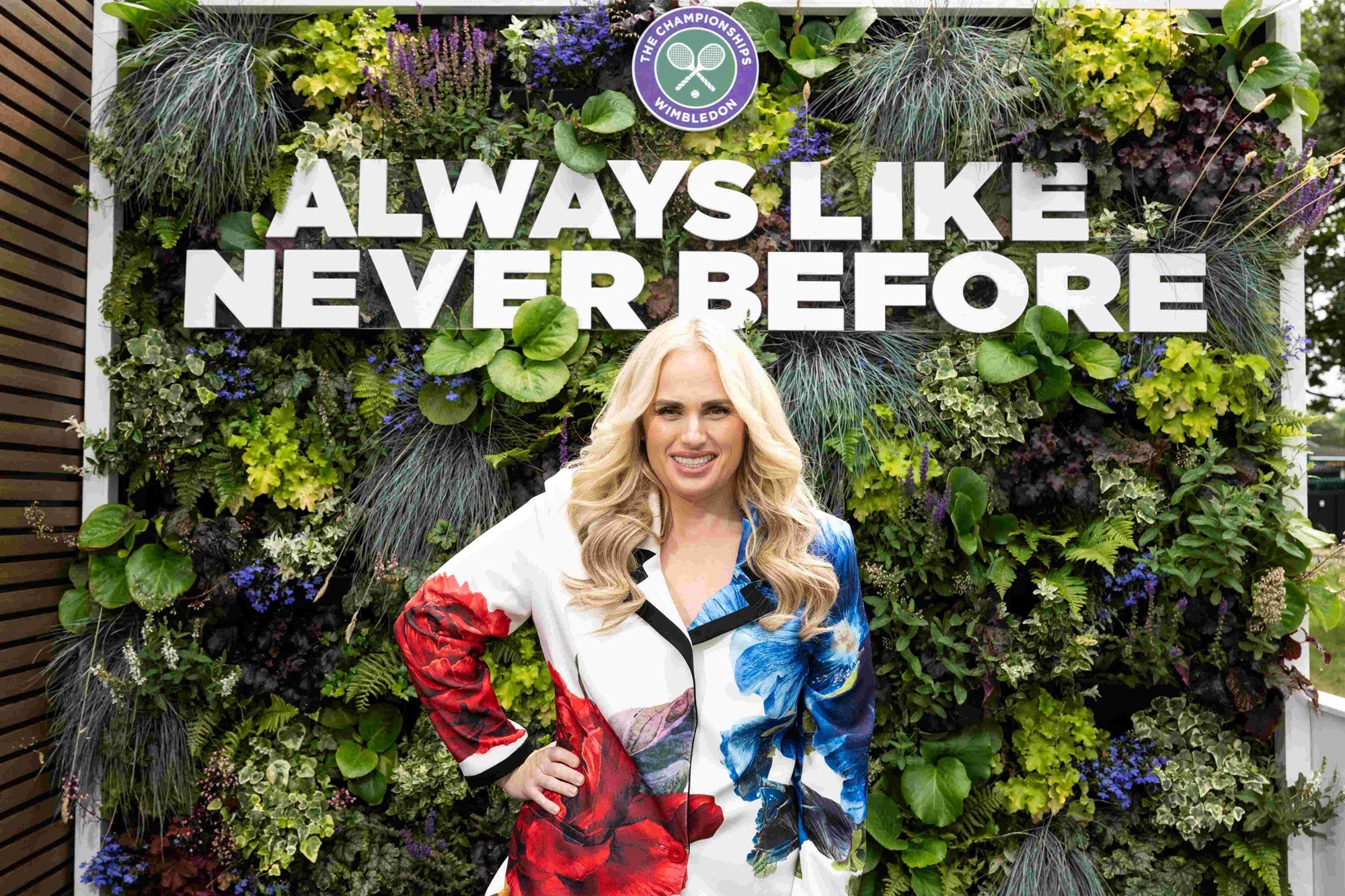 Rebel Wilson smiling in front of a wall of plants with the sign 'Always Like Never Before' hanging up