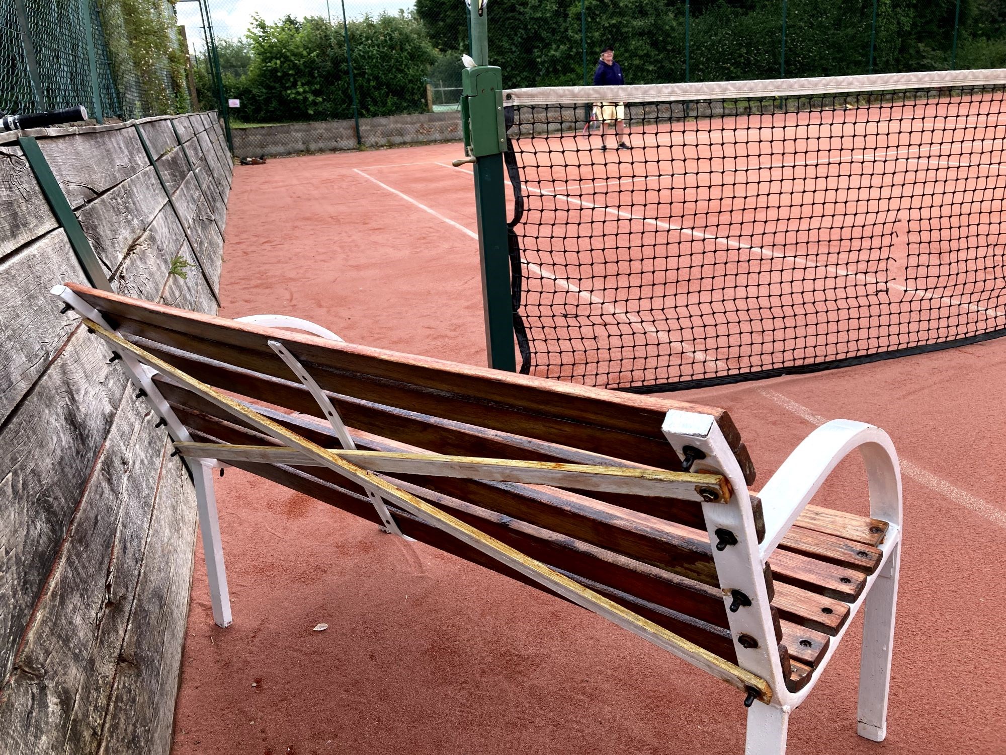 A bench with wooden struts attached diagonally across the back as a repair, situated on the side of a clay tennis court.