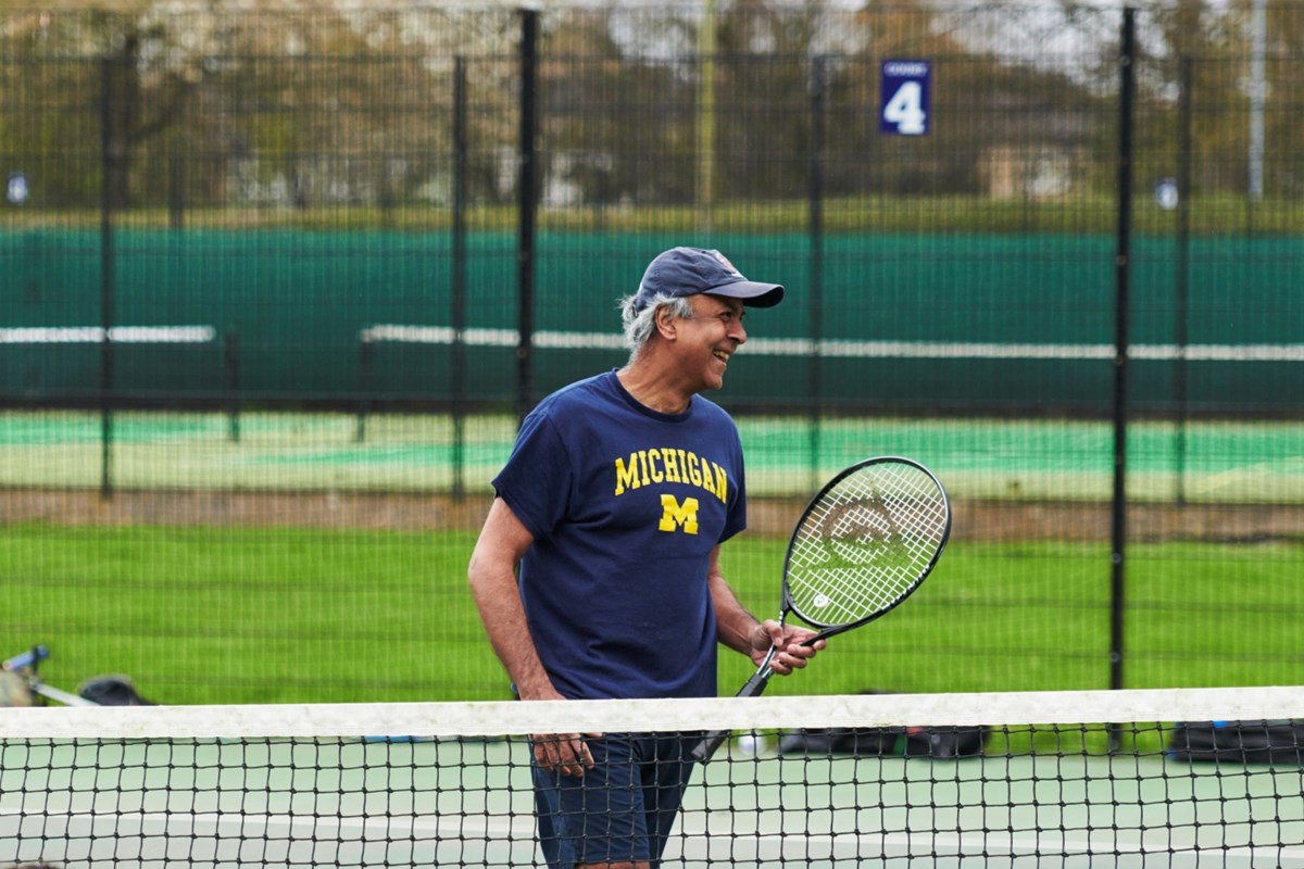 A man smiles while holding a tennis racket. He is stood behind a tennis net, wearing dark shorts, a dark blue t-shirt with the word 'Michigan' written in yellow font, and a grey/blue cap