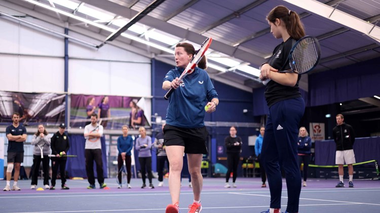A female coach stood on a tennis court holding a tennis racket and ball while a younger girl is stood next to her