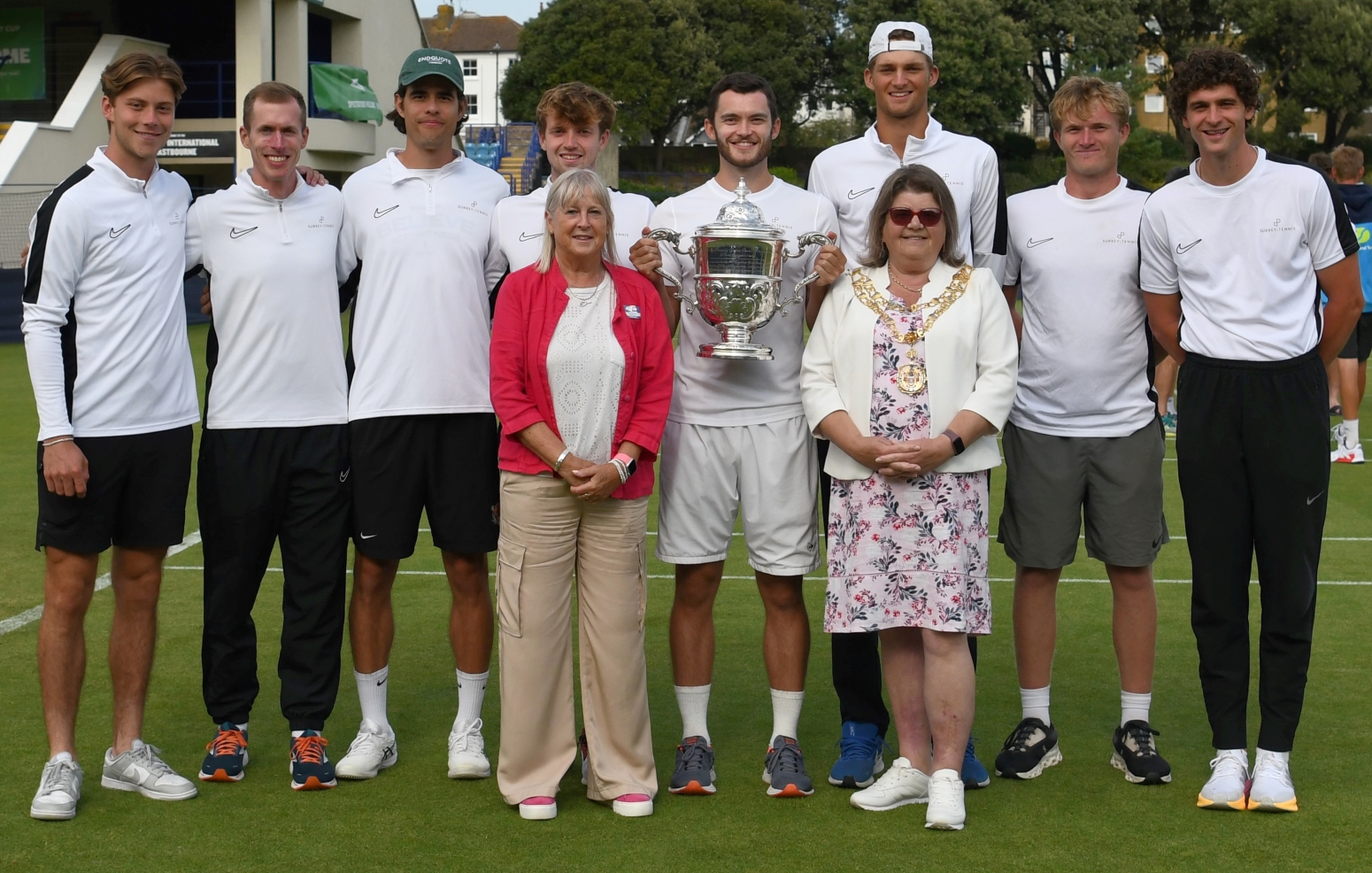 A group of men, all wearing white t-shirts and dark coloured shorts, stand on a grass tennis court with a trophy, with two women stood in the front either side of the trophy holder