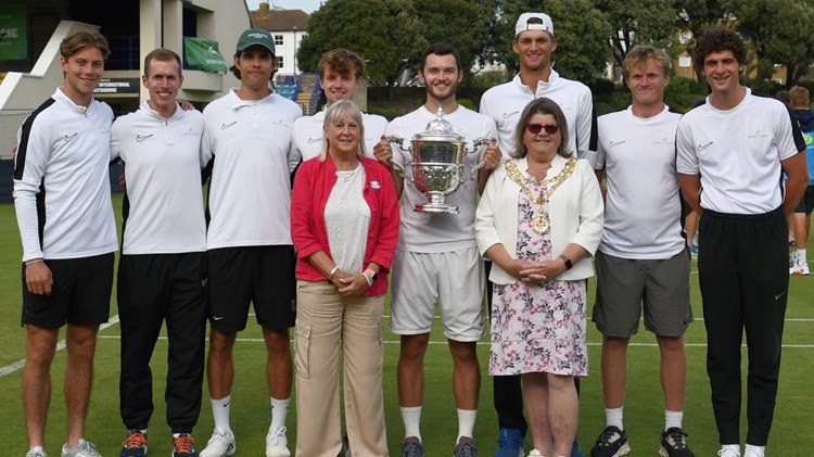 A group of men, all wearing white t-shirts and dark coloured shorts, stand on a grass tennis court with a trophy, with two women stood in the front either side of the trophy holder