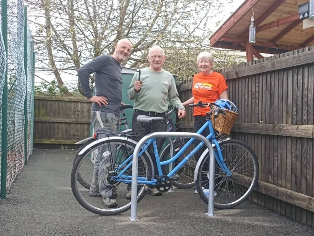 Three people, two men and a woman, stood behind a blue bicycle with a basket on the front containing a bike helmet