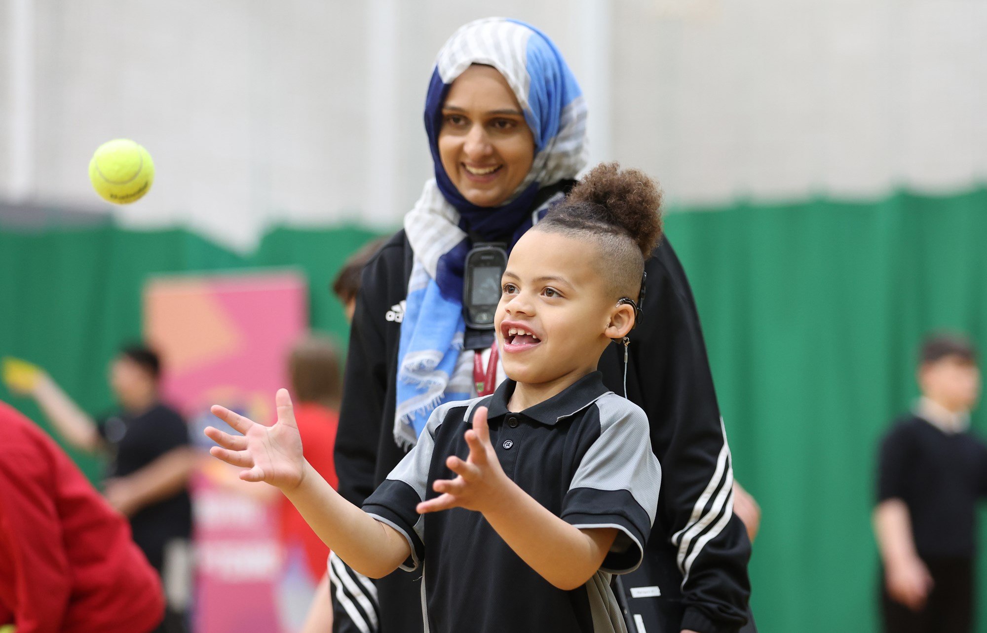 Young boy with a hearing aid, catching a tennis ball. A smiling coach is looking on.