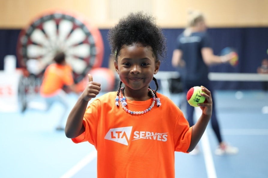 Young girl in an orange LTA SERVES t shirt holding a tennis ball in one hand and giving a tumbs up with the other