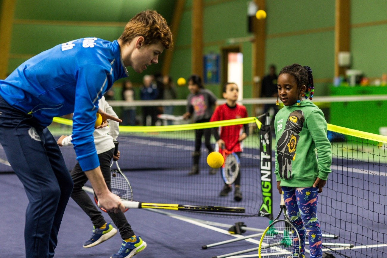 alt= An instructor in blue helps young children play tennis on an indoor court with mini nets and soft balls.