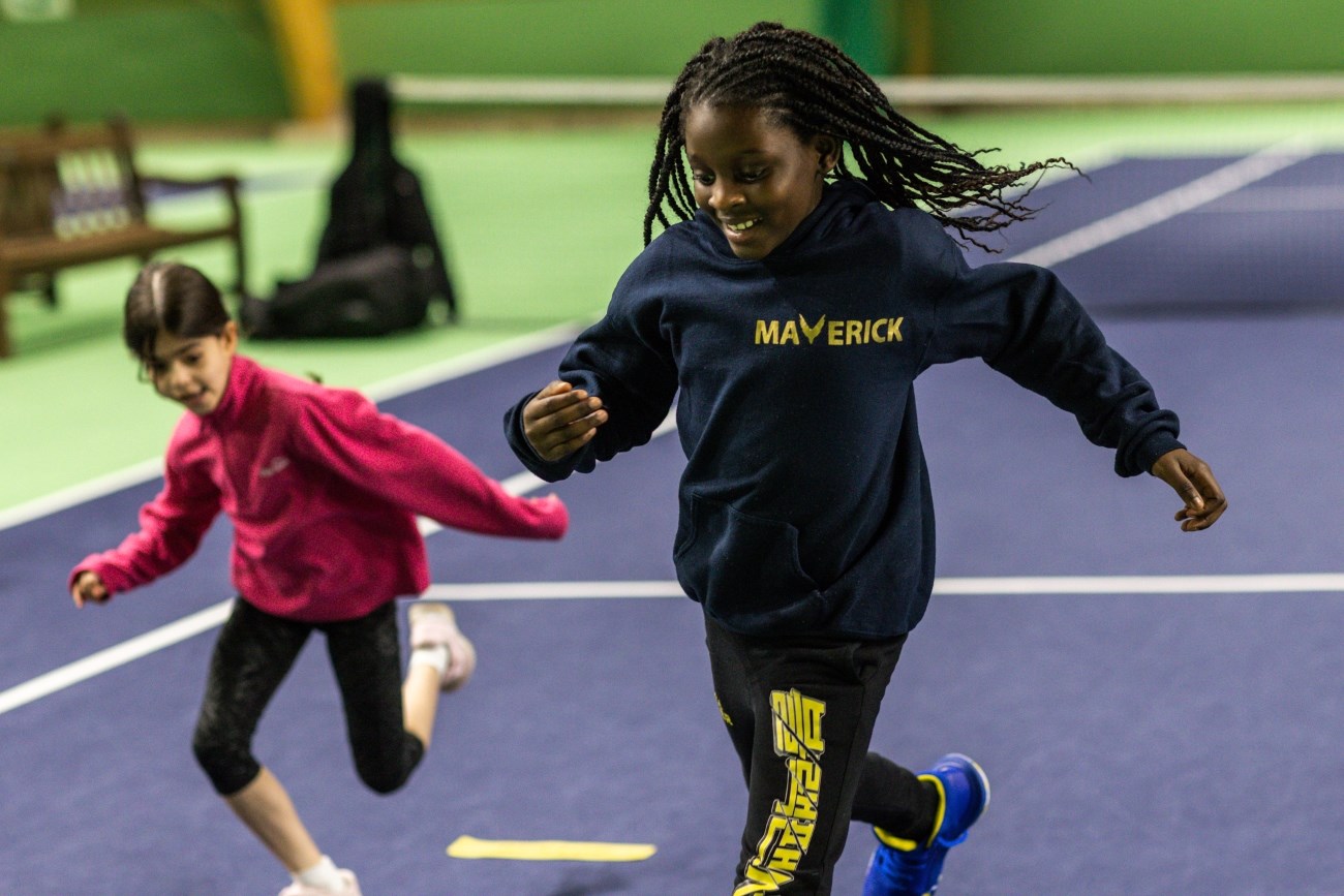 Two young girls running on an indoor tennis court. The girl in front is wearing a dark hoodie with the word 