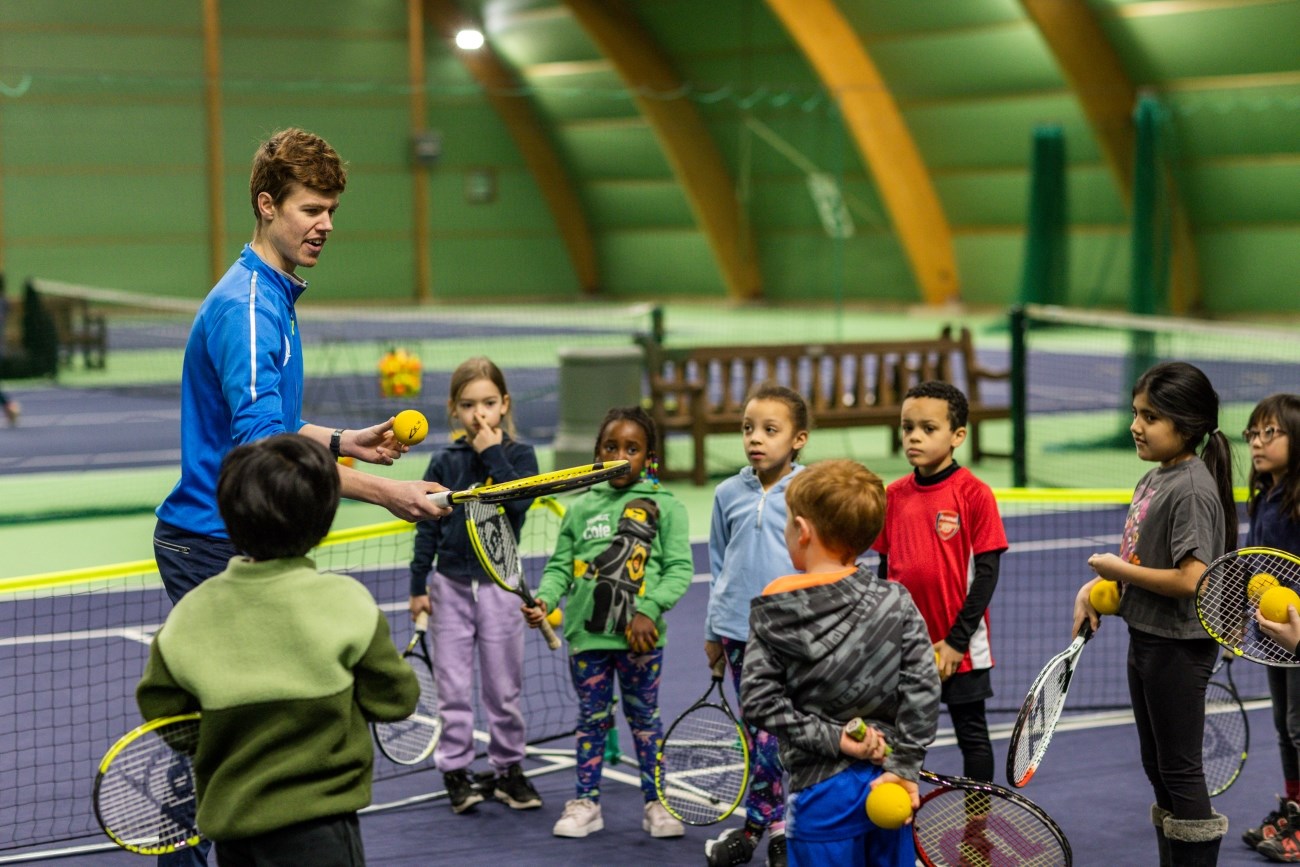 A tennis coach teaching a diverse group of children in an indoor tennis court. Kids hold rackets and tennis balls while listening attentively to the instructor.