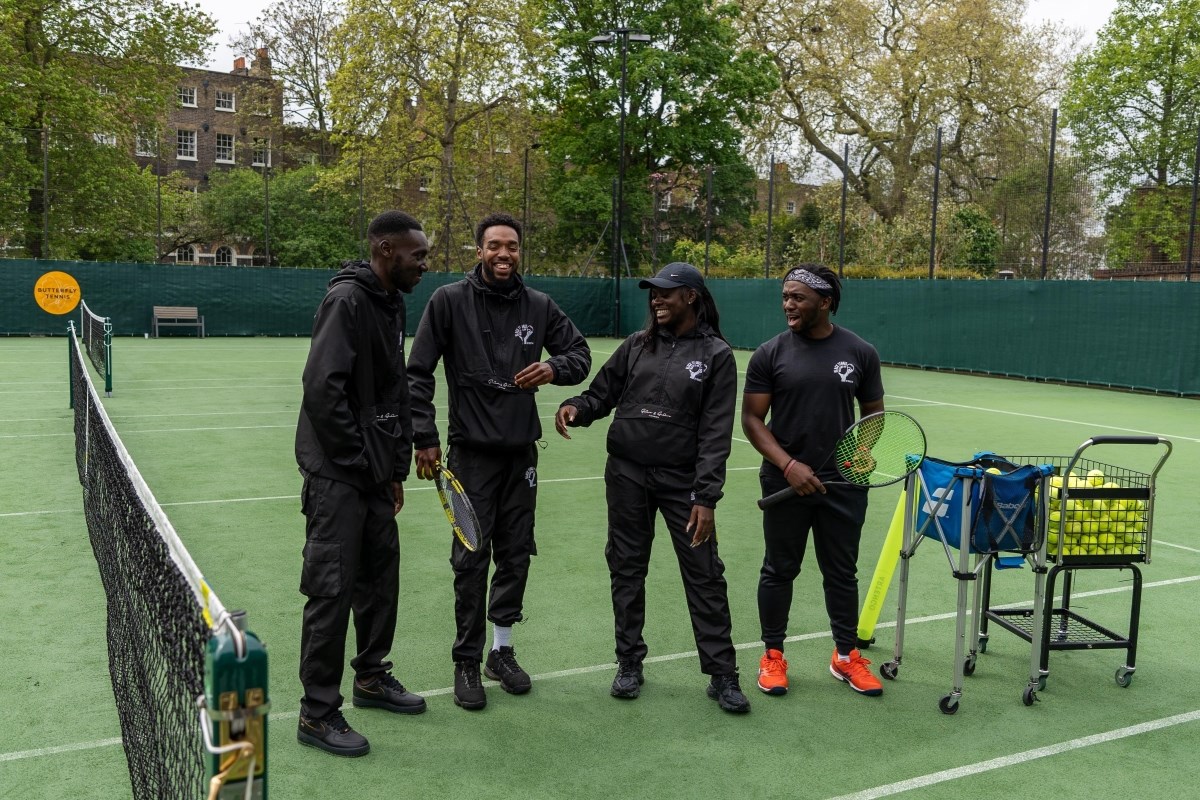 A group of four black people, all wearing black clothing, are stood together laughing on a tennis court, holding racquets and standing near a ball cart filled with tennis balls.