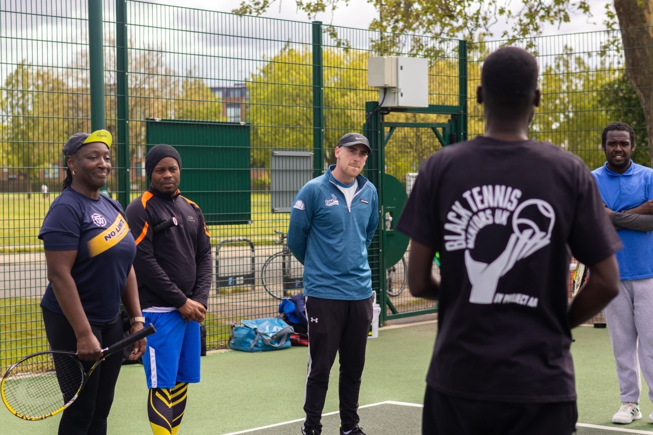 Diverse group of adults standing on an outdoor tennis court, attentively listening to a tennis coach. Trees and park visible in the background.