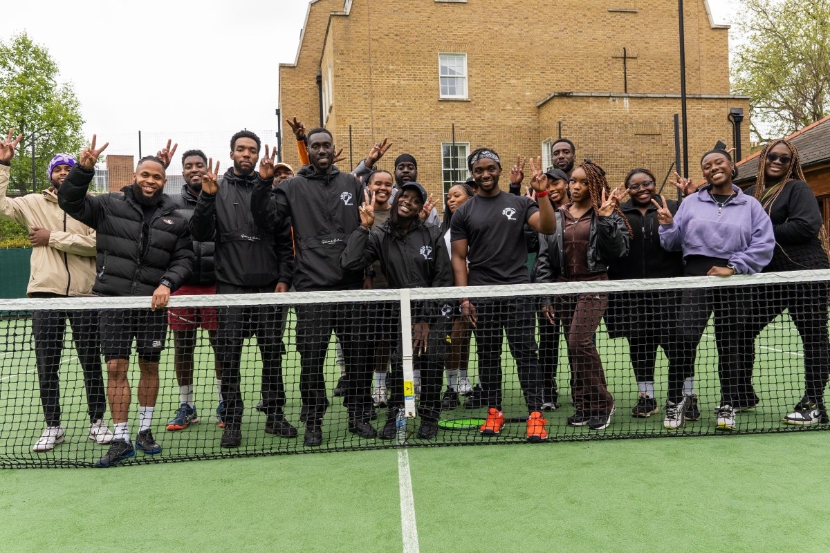 A group of young black adults posing on a tennis court, stood behind a net smiling and flashing peace signs. A large brick building is visible in the background.