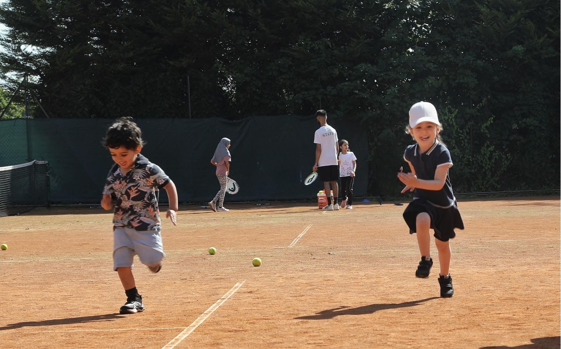 Two young children, a boy on the left and girl on the right, running on a clay tennis court. The girl is wearing a white cap. Tennis balls are on the ground behind them, while two other young children are receving coaching from an adult further in the background.