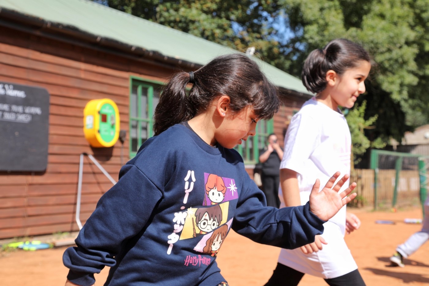 Two young girls are stood in running stances. The girl in the foreground is wearing a dark blue jumper with Japanese writing and animated characters on it, and is extending her arm in front of her as if running. The other girl in the background is wearing a white t-shirt and is stood with her arms by her side.