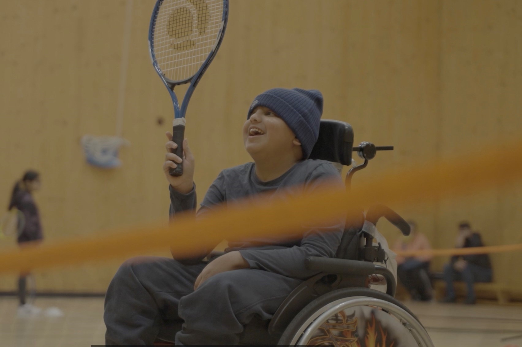 A boy sat in a wheelchair dressed in all-grey clothing, including a grey beanie hat, holds a tennis racket in the air in his right hand while smiling.