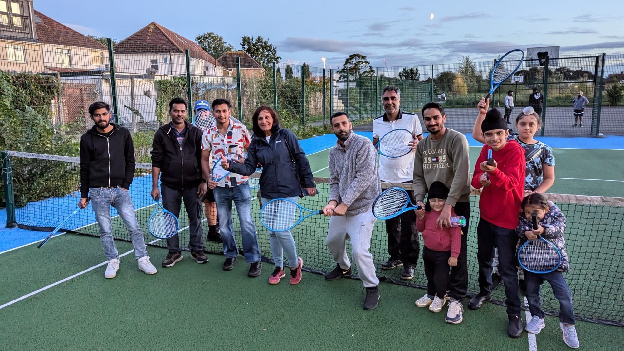 A group of adults and children holding their tennis rackets up in the air while stood next to a tennis net on a park court