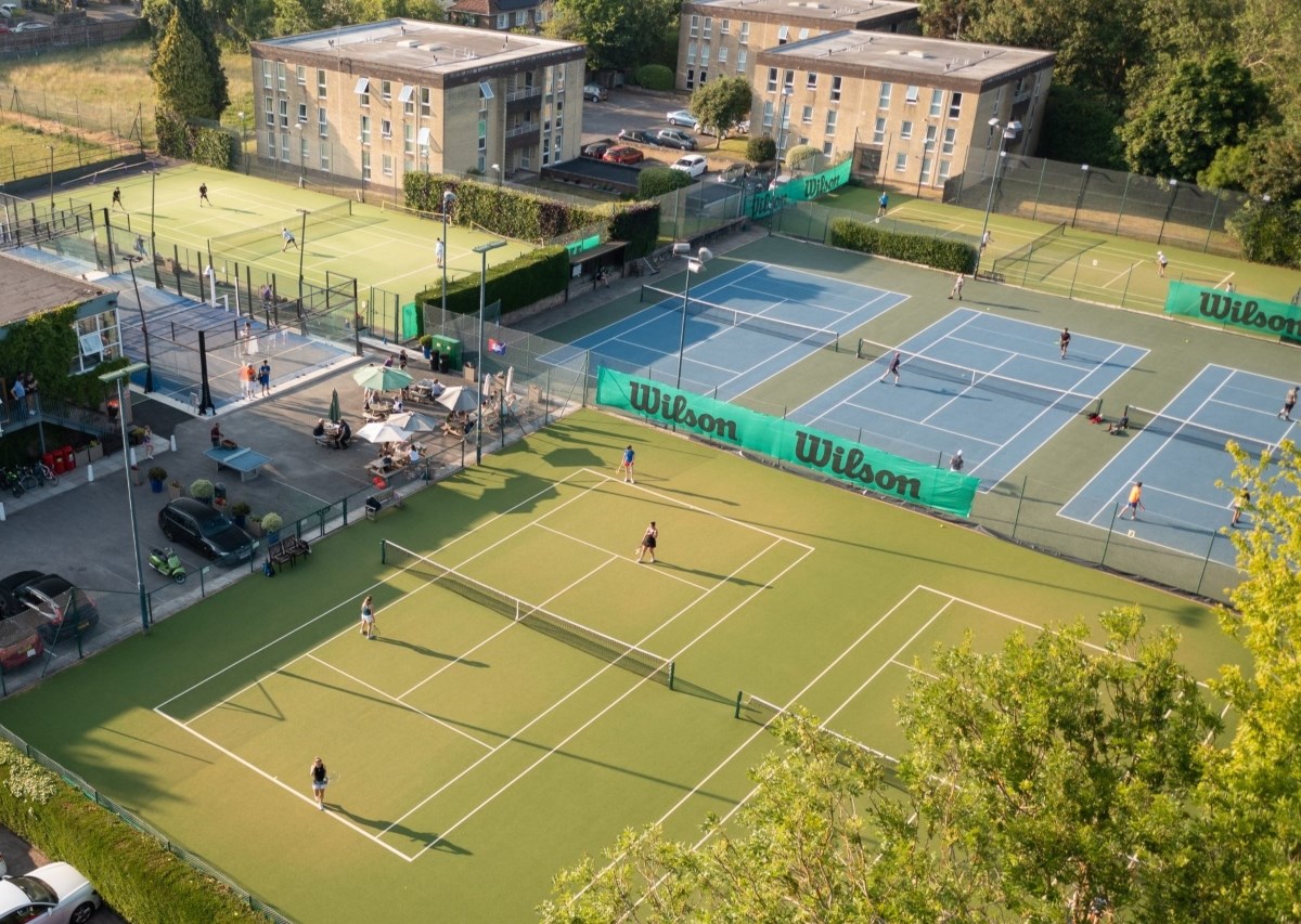 A wide view of green tennis courts with people playing on them, with one court in the foreground obscured by foliage from a large tree. In the background, there a a number of blocks of flats, in the bottom left corner, there is a white car parked behind a hedge. 